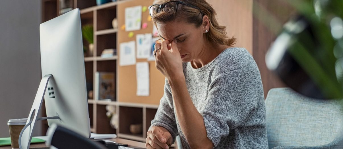 Exhausted businesswoman having a headache in modern office. Mature creative woman working at office desk with spectacles on head feeling tired. Stressed casual business woman feeling eye pain while overworking on desktop computer.