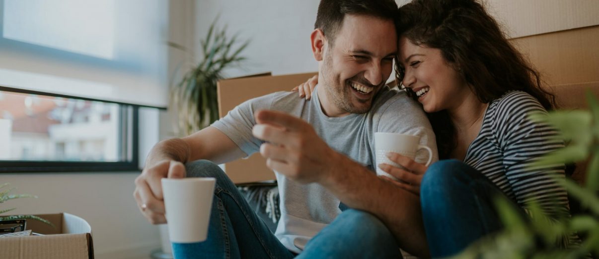 Young happy couple moving in new home and unpacking boxes. They are smiling and drinking a coffee.