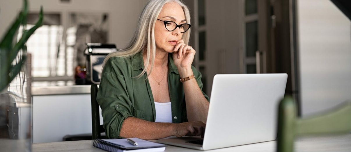 Focused old woman with white hair at home using laptop. Senior stylish entrepreneur with notebook and pen wearing eyeglasses working on computer at home. Serious woman analyzing and managing domestic bills and home finance.