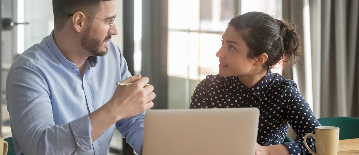 Caucasian mentor indian apprentice working together sitting at desk man explain work-related issues corporate goals give instructions, multi-ethnic co-workers share ideas information, teamwork concept