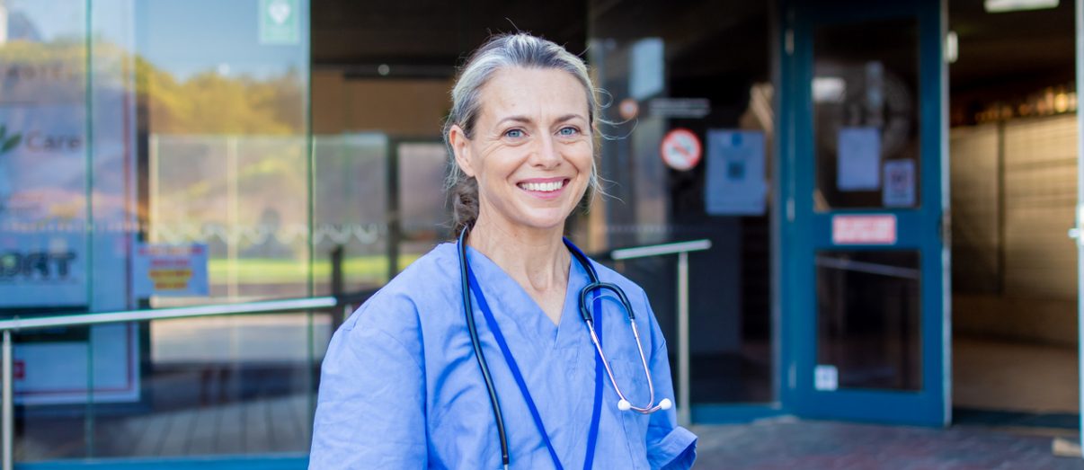 Aged Care 1 Smiling doctor wearing blue medical scrubs, standing in front of a hospital, looking directly at the camera and smiling.