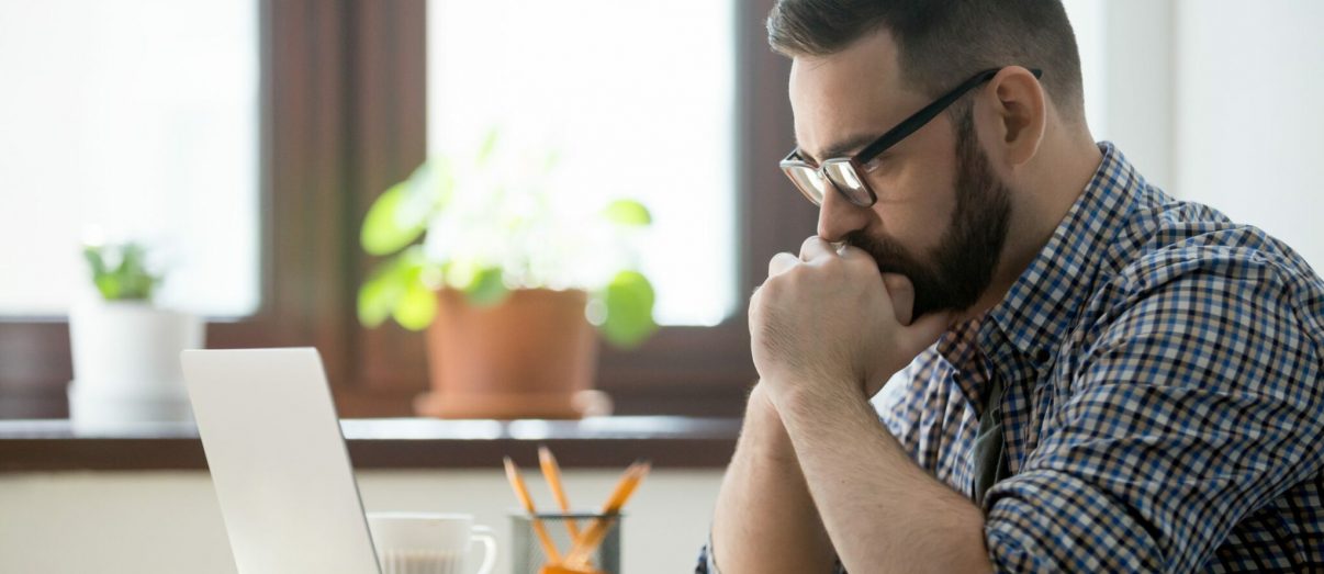 Young bearded manager working and reading data on laptop in home office. Thoughtful casual businessman thinking about job and looking on screen of notebook