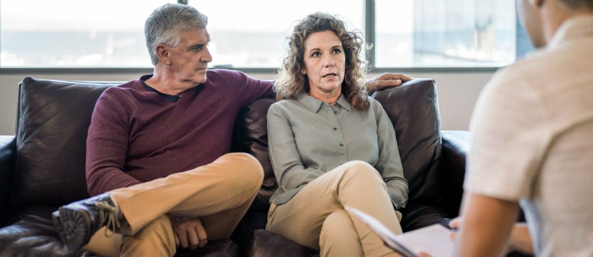 Mental health, financial and matrimonial professional with mature couple at office. Man is looking at woman sitting on psychiatrist's couch. They are wearing casuals.